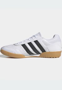 Chaussure de sport blanche avec des accents rayés noirs, des détails perforés, en maille textile, et une semelle en caoutchouc naturel, présentant un design bas.