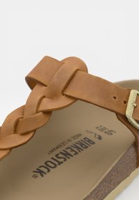 Rudi odos šlepetės su pintu dirželiu ir reguliuojamu sagtimi, ant pado išraižytas prekės ženklas "Birkenstock".