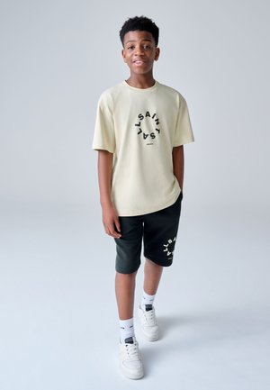 Dreng står og bærer beige oversized T-shirt og sorte shorts med cirkulært tekstdesign, hvide sneakers og sokker, mod en ensfarvet lys baggrund.