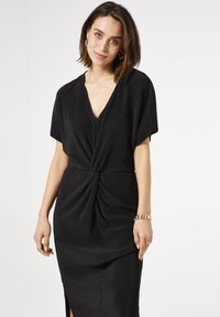 Vestido negro con un profundo escote en V, mangas cortas y un detalle de nudo retorcido en la cintura. Tejido liso y texturizado con aberturas laterales.