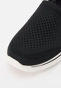 Sapatilha preta, tipo slip-on, com um upper em malha com um padrão texturizado, bico arredondado e sola em borracha branca com detalhes pretos.