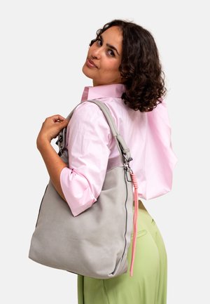 Graue Wildleder-Handtasche mit einer lässigen Form, die einen pinkfarbenen Akzent am Reißverschluss und schwarze Nähte aufweist. Eine Frau präsentiert die Tasche über der Schulter.