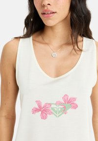 Weißes ärmelloses Oberteil mit V-Ausschnitt, versehen mit pinkfarbenen Hibiskusblüten und einem grünen Herz-Logo-Print auf der Vorderseite. Glatte Stofftextur.