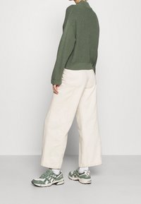 Sweter z dzianiny w oliwkowej zieleni, szerokie kremowe spodnie i zielono-białe buty sportowe. Sweter ma ściągacze na mankietach i dole oraz luźny krój.