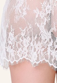 Nærbillede af hvidt blomstret blonderstof, der dækker huden, og viser indviklede net- og blomsterudskæringer med bølgede kanter.