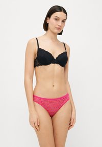 Schwarzer Spitzen-BH mit vorgeformten Cups und gewellten Kanten, kombiniert mit pinkfarbenen Netzslips mit kreisförmigem Muster. Beide Teile sind leicht.