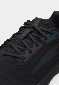 Μαύρο αθλητικό παπούτσι με επάνω μέρος από διχτυωτό ύφασμα, μαύρα κορδόνια και ιριδίζοντα διακοσμητικά κοντά στις τρύπες των κορδονιών και στο πλευρικό λογότυπο, κοντινή όψη.