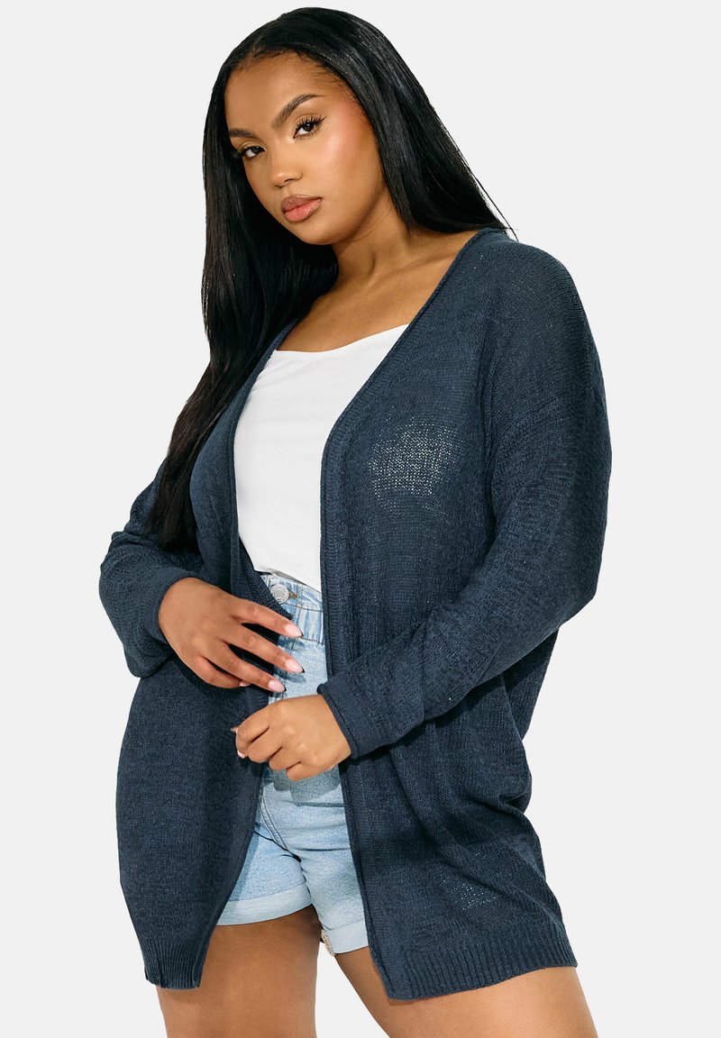 Marinblå cardigan i texturerad stickad tyg, öppen frontdesign, långa ärmar och avslappnad passform, i kombination med en vit topp och denimshorts.