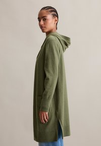 Cardigan en tricot vert olive avec capuche, présentant une fente latérale et une poche avant. Manches longues avec poignets côtelés. Texture douce.