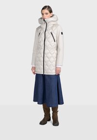 Prešívaná puffer bunda v svetlosivej farbe s vysokým golierom, bočnými zipsami a priliehavým strihom, spojená s tmavými riflovými sukňami a vysokými hnedými čižmami.