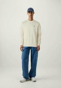 Suéter creme, boné azul, calças cargo soltas azuis, ténis brancos. O suéter tem um pequeno logótipo no peito, ajuste casual, tecido texturizado.
