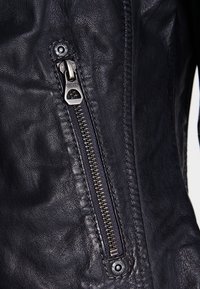 Zwarte leren jack met een opvallende rits met een zilveren trekband. Getextureerde afwerking en zichtbare stikdetails.