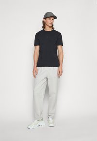 T-shirt preta de mangas curtas, calças beje claras e boné cinza. Calçado são ténis grossos brancos com detalhes verdes claros.