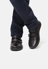 Black leather shoes with two adjustable straps, rounded toes, and a textured finish, paired with dark blue denim jeans.
