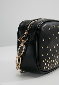 Černá kožená taška s zlatými cvočky a zipovým uzávěrem. Zlatý řetězový prvek se k tašce připojuje pro zvýšení detailu.