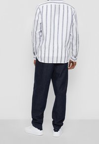 Camicia in lino bianco con righe verticali blu scuro, vestibilità ampia, orlo curvo. Abbinata a pantaloni in denim scuro e sneakers bianche.