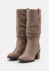 Bottes en suède marron montant jusqu'au genou avec un design légèrement ample, bout pointu et talon empilé. Présente un petit détail en boucle sur le côté.