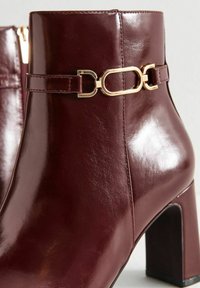 Botas de tobillo de cuero burdeos con un acabado brillante, presentan un detalle de cadena dorada y un tacón cuadrado para mayor estabilidad. Cierre de cremallera en la parte trasera.