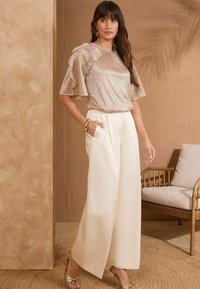 Top in bronzo scintillante con maniche a volant, abbinato a pantaloni bianchi larghi. Tacchi con accenti dorati completano l'outfit. Sfondo neutro.