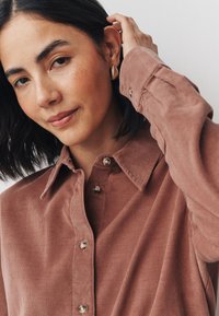 Porcelánrózsaszín kordbársony pomponos galléros ing, gombos záródással és texturált anyaggal. A modell egy fülbevalót visel, miközben a haját igazítja.