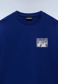 Tummansininen puuvillapaita, jossa on pyöreä pääntie. Paidassa on vasemmassa rinnassa valkoinen graafinen logo-patch, jossa on palmu. 