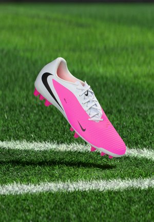 Chaussure de football Nike blanche et rose avec un swoosh noir flottant au-dessus de l'herbe verte près d'une ligne blanche du terrain.
