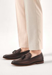 Hnedé kožené mokasíny s strapcami, s okrúhlou špičkou a kontrastnou hnedou podrážkou. Kombinované s ľahkými béžovými nohavicami na mieru.