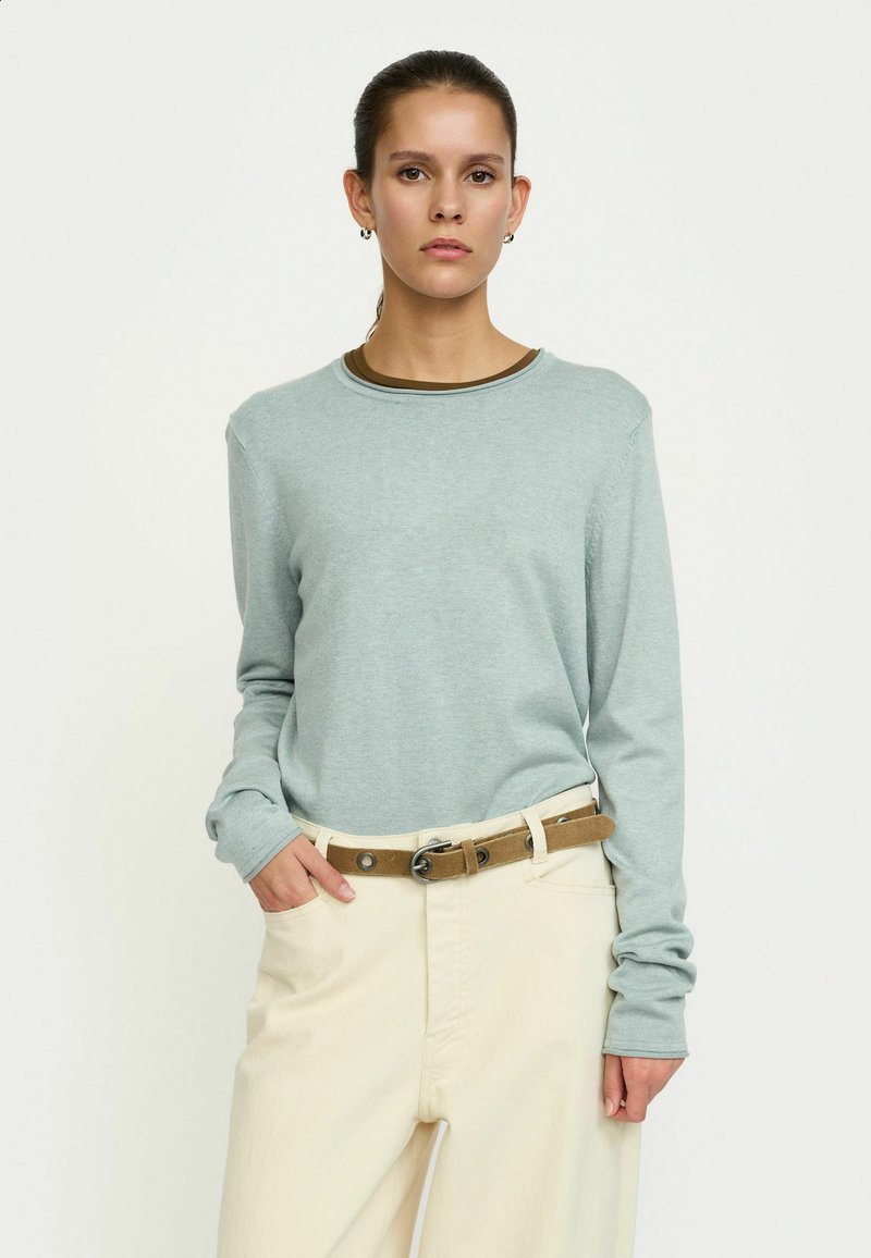 Chemise bleu clair à manches longues avec un col rond et un tissu légèrement texturé, associée à un pantalon de couleur crème et une ceinture marron.