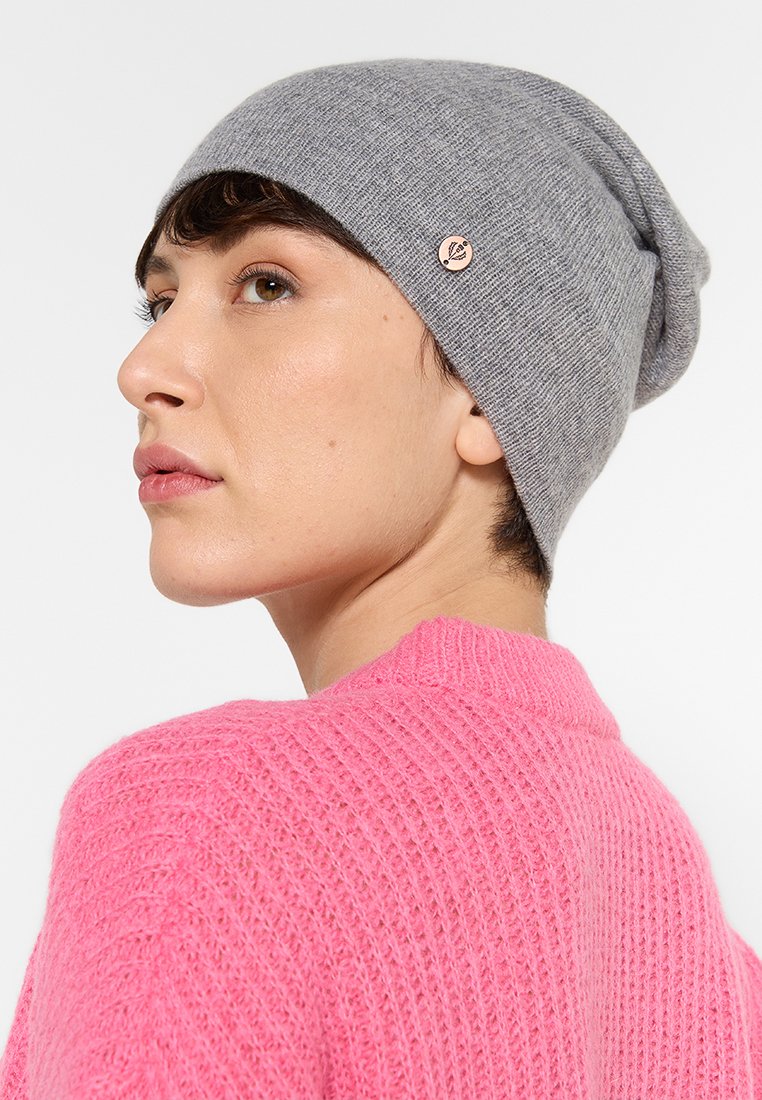 Młoda osoba z krótkimi ciemnymi włosami, nosząca szary dzianinowy beret i jasnoróżowy sweter o fakturze, patrząca lekko w górę i w lewo.