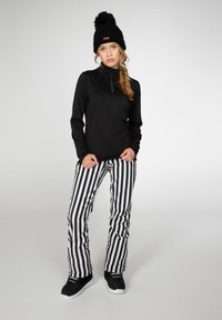 Blusa negra de manga larga con cuello de media cremallera, combinada con pantalones a rayas verticales en negro y blanco. Gorro negro con pom-pom en la cabeza.