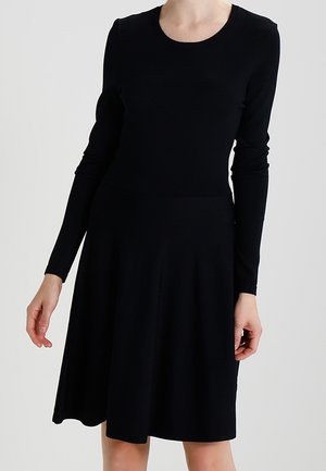 Frau trägt ein schlichtes schwarzes, knielanges Kleid mit langen Ärmeln und steht vor einem hellgrauen Hintergrund.