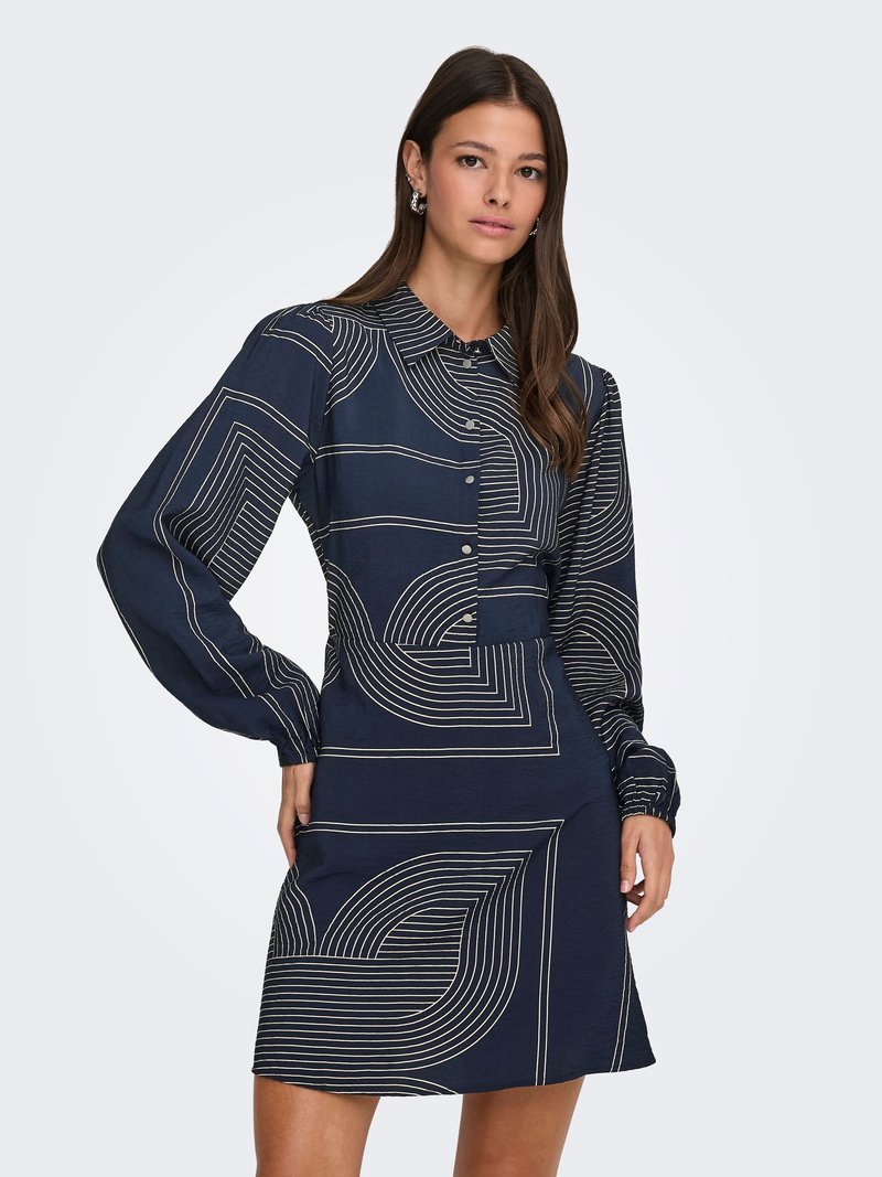 Frau trägt ein dunkelblaues Kleid mit weißen geometrischen Linienmustern, langen Ärmeln, einem Kragen und Knöpfen vorne, steht vor einem einfachen hellen Hintergrund.