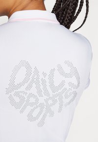 Tricou polo alb cu mânecă scurtă, prezentând un design strălucitor din strasuri pe spate care scrie "Daisy Sports" cu un font jucăuș.