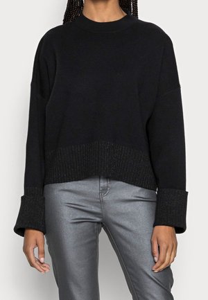 Personne portant un pull noir à manches longues avec des poignets larges et un pantalon argenté brillant, debout avec les bras détendus le long du corps.