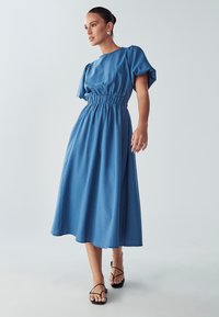 Modra midi obleka z zbranimi bokovi, kratkimi puff rokavi in tekočim robom. Oblečena z črnimi sandali s trakovi. Gladka tekstura tkanine.