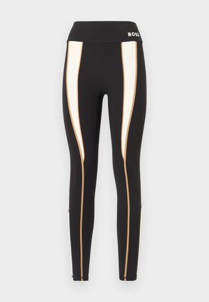 Črne legice z visokim pasom, ki imajo bele in zlate vertikalne črte ob straneh ter gladek, raztegljiv material.