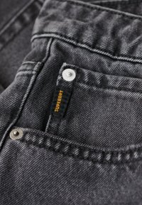 Jean en denim gris foncé avec une étiquette de logo Superdry bien visible, des boutons en métal et des coutures contrastantes le long des poches.