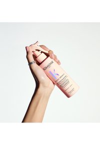 Kérastase Gloss Absolu fles in zacht roze met een spraymond, voorzien van zwarte tekst en holografische accenten, vastgehouden in een hand tegen een witte achtergrond.