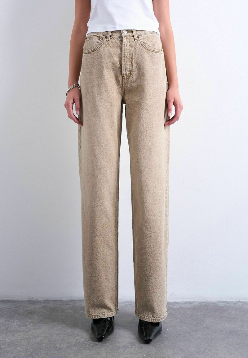 Persoon die beige, rechte, high-waist jeans draagt met een witte top en zwarte puntige schoenen, staand tegen een effen achtergrond.