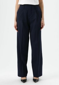 Pantalon taille haute bleu marine avec plis, en tissu lisse. Caractéristiques jambes larges et coupe ajustée, associé à des chaussures noires pointues.