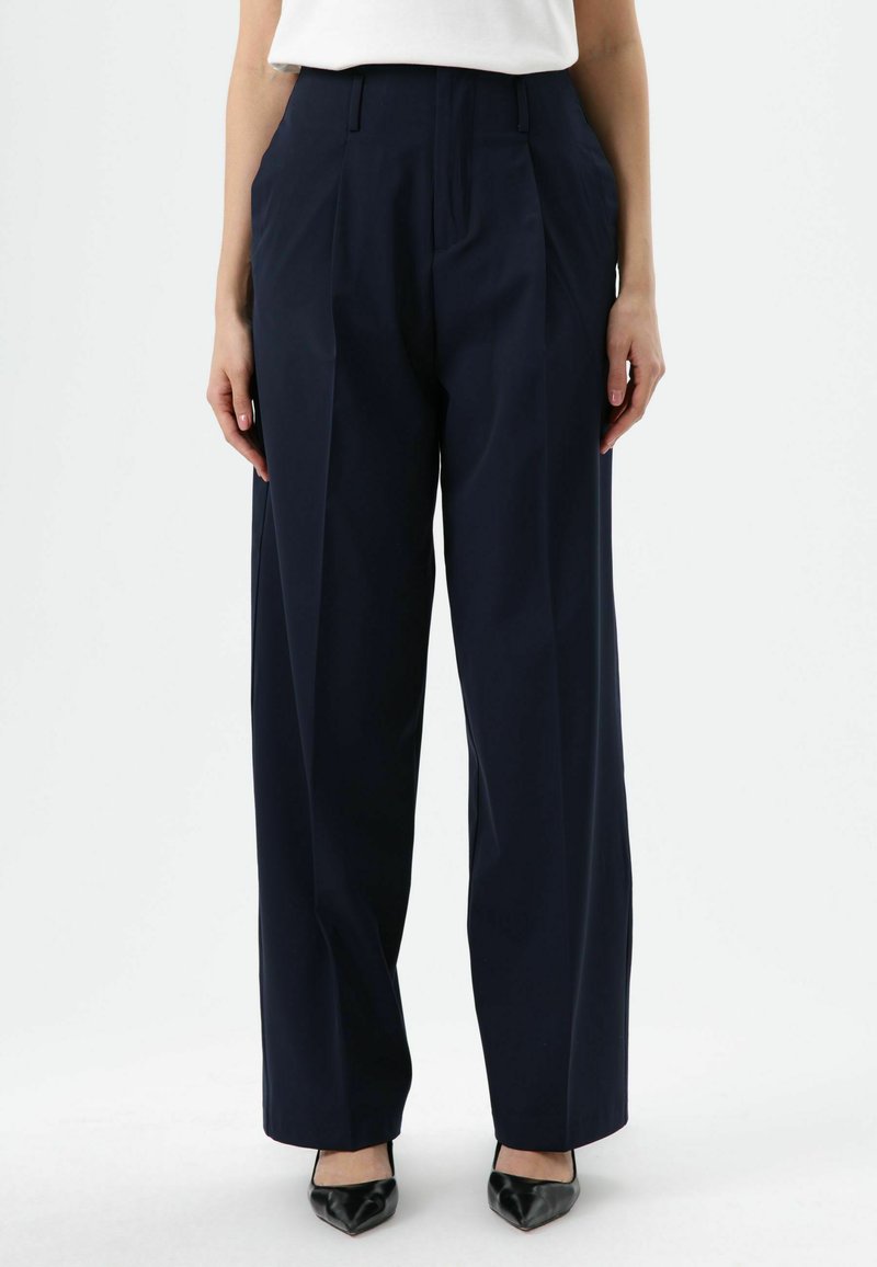 Pantalon taille haute bleu marine avec plis, en tissu lisse. Caractéristiques jambes larges et coupe ajustée, associé à des chaussures noires pointues.