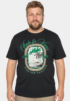 Uomo barbuto che indossa una maglietta nera con un motivo tropicale verde e beige con palme, onde e testo "Made for Waves Aloha Born for Paradise."