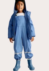 Blå vandtæt jakke med hætte, matchende overalls med en lomme foran og reflekterende stribe, båret over blå gummistøvler.