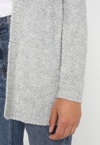 Cardigan lavorato a maglia grigio chiaro con finitura testurizzata e polsini a coste, indossato sopra dei jeans, che mostra una vestibilità ampia e un design davanti aperto.