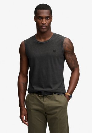 Hombre con pelo corto que lleva una camiseta negra sin mangas, pantalones oliva, cinturón negro, reloj de pulsera y tatuajes visibles en el brazo derecho.