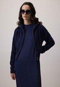 Tumši zila zipa jaka ar teksturētu audumu un ribotām aprocēm, uzvilkta virs vienkrāsaina tumši zilā kleita, kombinēta ar lielām tumšām saules brillēm.