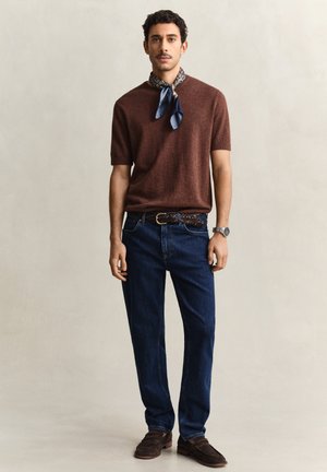 Homme debout portant un pull à manches courtes marron, une écharpe bleue à motifs autour du cou, un jean bleu foncé avec une ceinture tressée, et des mocassins marron.