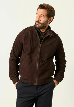 Mann mit Bart, der eine dunkelbraune Wildleder-Zipjacke und schwarze Hose trägt, schaut seitlich mit den Händen in den Taschen vor einem einfarbigen Hintergrund.