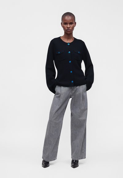 Personne portant un cardigan noir avec des boutons bleus, un pantalon gris à jambes larges et des chaussures noires pointues, debout les mains dans les poches devant un fond uni.