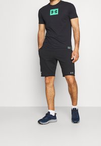 Svart kortärmad t-shirt med en grön Under Armour-logga, i kombination med svarta shorts och marinblå träningsskor, med vita strumpor.